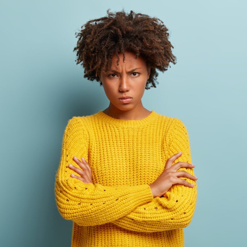 Annoyed pissed woman crosses hands over chest, frowns and sulks, waits for explanation from boyfriend, looks from eyebrows, wears knitted yellow sweater, stands against blue wall, feels insulted